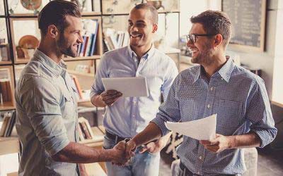 Three men shaking hands in a casual indoor setting, with documents in their hands.