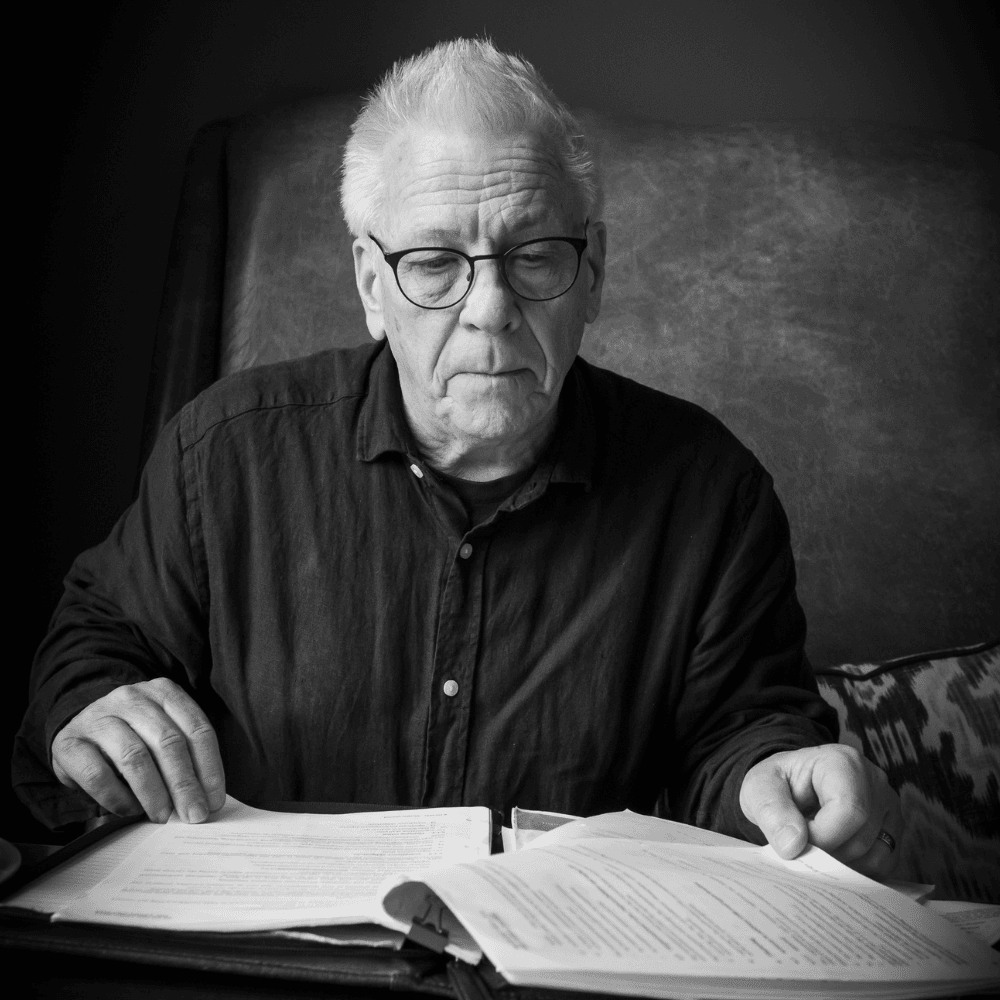 Older man with glasses reading documents while seated, wearing a black shirt.